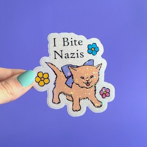 May include: A sticker featuring a cartoon kitten with a blue bow, surrounded by flowers. The text "I Bite Nazis" is above the kitten. The sticker has a holographic, glittery effect and is on a purple background.