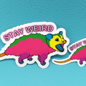 May include: Two colorful opossum stickers with the text "STAY WEIRD". The opossums are depicted in bright pink, green, yellow, and blue. One sticker is larger than the other. The stickers are on a light blue background.