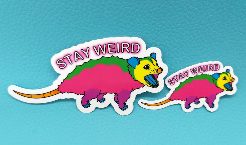 May include: Two colorful opossum stickers with the text "STAY WEIRD". The opossums are depicted in bright pink, green, yellow, and blue. One sticker is larger than the other. The stickers are on a light blue background.