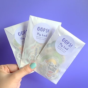 May include: Three white envelopes with a sticker on each. The stickers are round and have the text "OOPS! My bad Waterproof vinyl B grade stickers" in purple text.