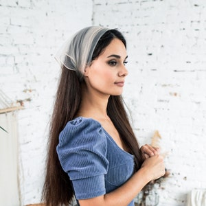 Pu&ograve; includere: Una donna con lunghi capelli castani indossa un abito di velluto a coste blu chiaro con maniche a sbuffo e un foulard bianco trasparente.