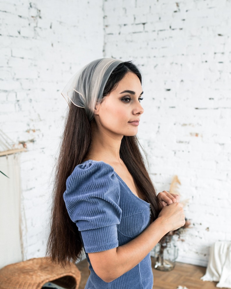 Pu&ograve; includere: Una donna con lunghi capelli castani indossa un abito di velluto a coste blu chiaro con maniche a sbuffo e un foulard bianco trasparente.