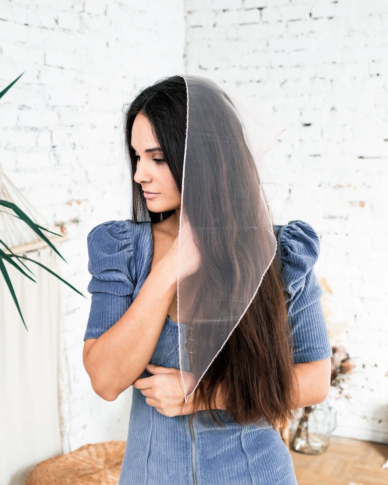 May include: A woman with long dark hair wearing a blue corduroy dress and a sheer pink scarf. The scarf is draped over her head and shoulders.