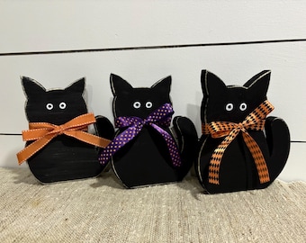 May include: Three black wooden cat figurines with white eyes. Each cat is wearing a different colored bow. The bows are orange with white polka dots, purple with white polka dots, and orange and black checkered.