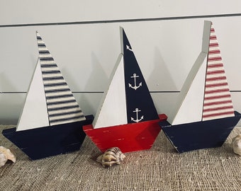 May include: Three wooden sailboat decorations with navy blue hulls and white sails. One sail has navy blue and white stripes, another is red with white anchors, and the third has red and white stripes. Seashells are placed in front of the boats.