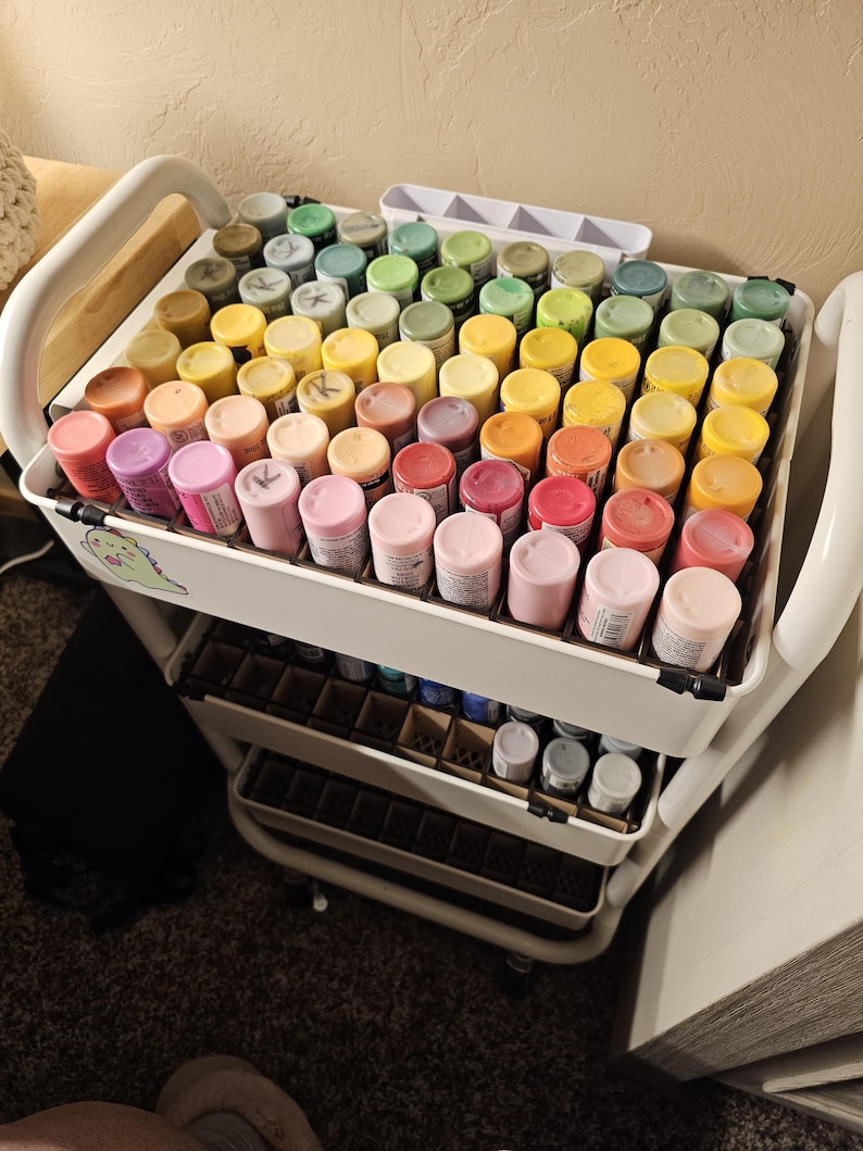 May include: A white rolling cart with three shelves, each filled with small bottles of paint. The bottles are arranged in rows by color, starting with green, yellow, orange, red, pink, and ending with purple. The cart has a handle on the side and is on a carpeted floor.