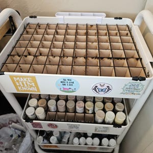 May include: White rolling cart with three shelves. The top shelf has a gridded organizer with 36 compartments. The middle shelf has 16 small bottles in a gridded organizer. The bottom shelf has 12 small bottles in a gridded organizer. The cart has stickers with the text "Make Him Known", "All You Need is Love and a Dog", "Joy & Peace", and a butterfly with a crescent moon.