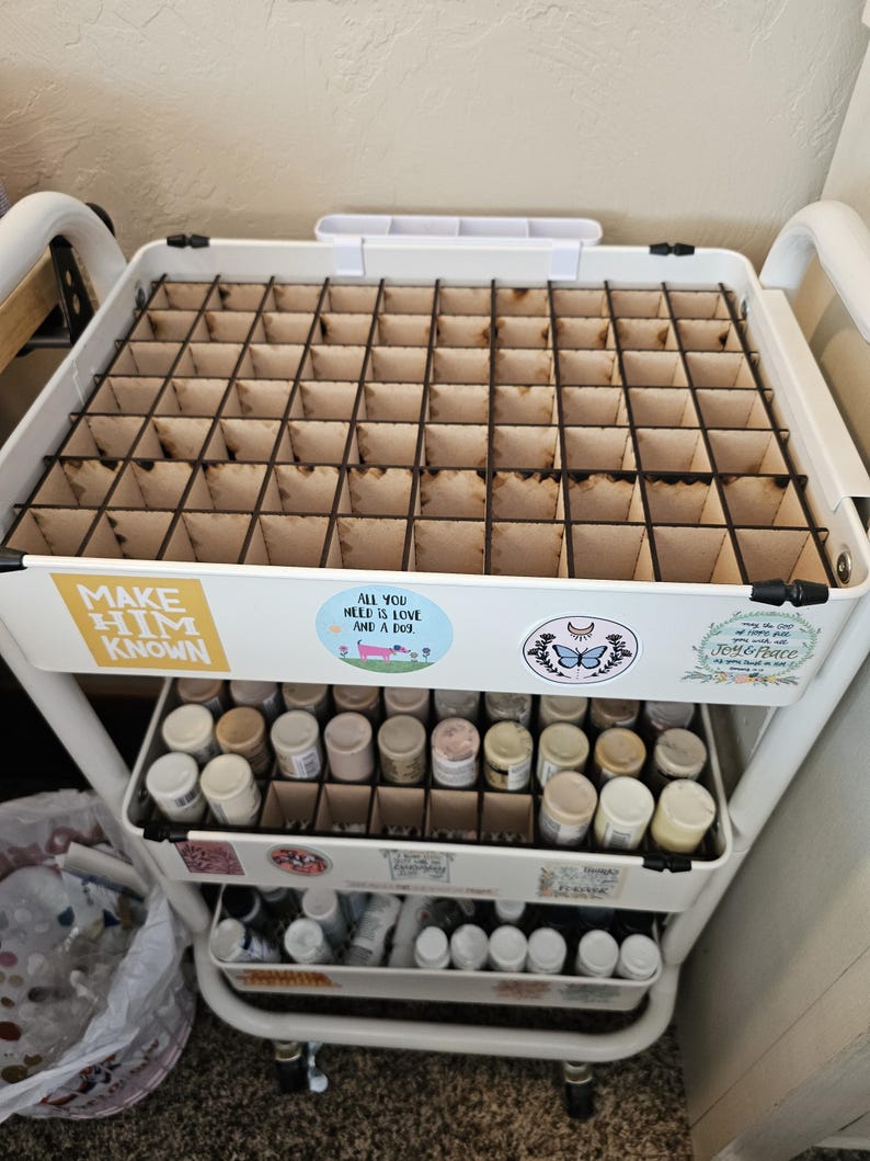 May include: White rolling cart with three shelves. The top shelf has a gridded organizer with 36 compartments. The middle shelf has 16 small bottles in a gridded organizer. The bottom shelf has 12 small bottles in a gridded organizer. The cart has stickers with the text "Make Him Known", "All You Need is Love and a Dog", "Joy & Peace", and a butterfly with a crescent moon.