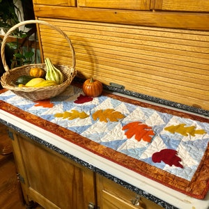 May include: A blue and white quilted table runner with a brown, orange, and red leaf design. The runner is on a wooden countertop with a basket of gourds and a small pumpkin.