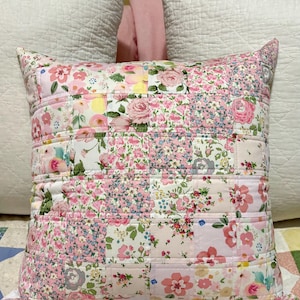 May include: A pink and white floral patchwork cushion with a quilted design. The cushion is made of various floral fabrics sewn together in a patchwork pattern.