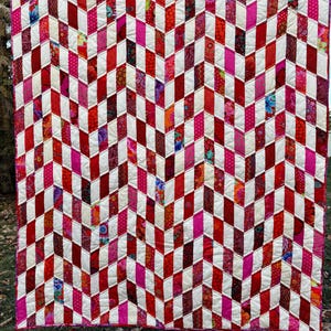 May include: A handmade quilt with a geometric chevron pattern. The quilt features alternating rows of red, white, and pink fabric rectangles. Some rectangles have floral and patterned designs. The quilt is hanging outside on a washing line.