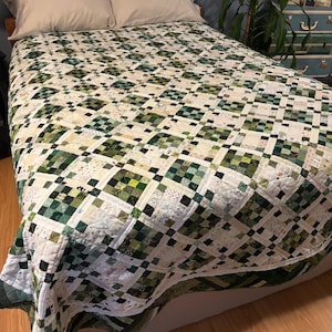 May include: A handmade quilt with a patchwork design in shades of green, white, and black covers a bed. The quilt features a repeating square pattern. Two beige pillows rest on the bed. The quilt is likely queen or king size.