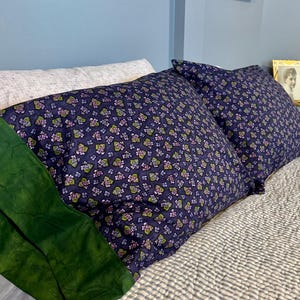 May include: Two navy blue pillowcases with a floral pattern of pink, purple, and green flowers. The pillows are on a bed with a blue and white striped quilt. A green fabric and a white pillow are also visible.