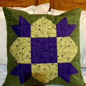 May include: A green and purple quilted cushion with a starburst design. The cushion is made of fabric with a floral pattern and has a quilted design.