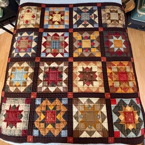 May include: A patchwork quilt with a repeating pattern of square blocks. Each block features a star or geometric design in various colors, including red, blue, yellow, and brown. The quilt is made of fabric squares and has a dark brown border.