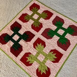 Op de afbeelding: Handgemaakte quilt met een patroon van rode aardbeien met groene stelen en bladeren. De quilt heeft een lichtroze achtergrond met kleine witte stippen en een groene rand. Geplaatst op een gestreept, lichtblauw en wit sprei.