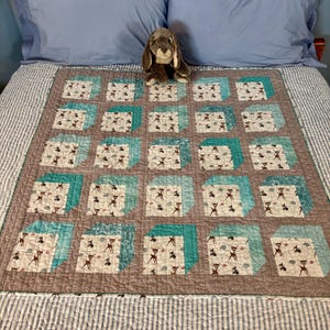May include: A handmade quilt with a deer pattern in a grid design. The quilt features squares with deer and cloud motifs, framed by teal and brown borders. A stuffed bunny sits on top of the quilt, which is placed on a bed.