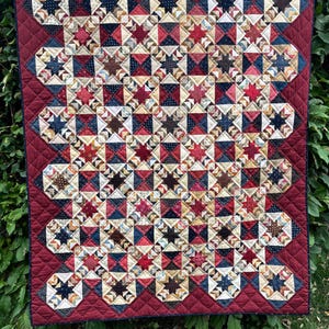 May include: A rectangular quilt featuring a repeating star pattern in shades of red, navy blue, cream, and brown. The quilt has a solid deep red border with a diamond-shaped quilted texture and is constructed from various patterned fabric squares.