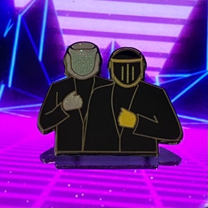 Puede incluir: Un pin de esmalte negro y dorado con dos figuras que llevan cascos. La figura de la izquierda lleva un casco blanco con textura brillante. La figura de la derecha lleva un casco negro con detalles dorados y una serie de líneas verticales.