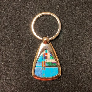 May include: A silver-toned keychain with a teardrop-shaped pendant. The pendant features a mosaic design with turquoise, blue, red, and white geometric shapes. The keychain has a round ring for attaching keys.