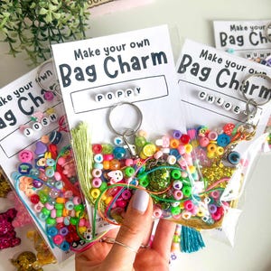 May include: A clear plastic bag containing colorful beads, a keychain, and a tassel. The bag has a white label that reads "Make your own Bag Charm" with the name "POPPY" spelled out. Other bags are visible with the names "JOSH" and "CHLOE".