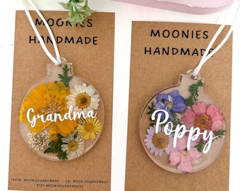 May include: Two round, clear resin ornaments with pressed flowers and a white name. The ornament on the left has yellow and white flowers and the name "Grandma". The ornament on the right has pink, purple, and blue flowers and the name "Poppy".
