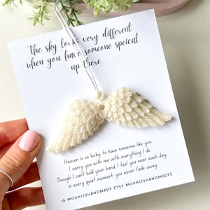 May include: A cream-coloured angel wing ornament with a white card featuring handwritten text. The text includes phrases like "the sky looks very different" and "Heaven is so lucky." The ornament is suspended by a clear string.
