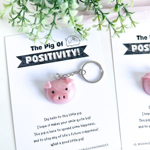 May include: A pink pig-shaped keychain with a silver chain and keyring, displayed on a card with the text "The Pig Of POSITIVITY!". The card also includes a message about spreading happiness. The background includes greenery.