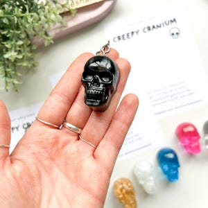 May include: A black skull-shaped keychain is held in a hand, with a silver ring attached. The keychain is glossy and detailed. The text "CREEPY CRANIUM" is visible on a card in the background, along with other colorful skull charms.