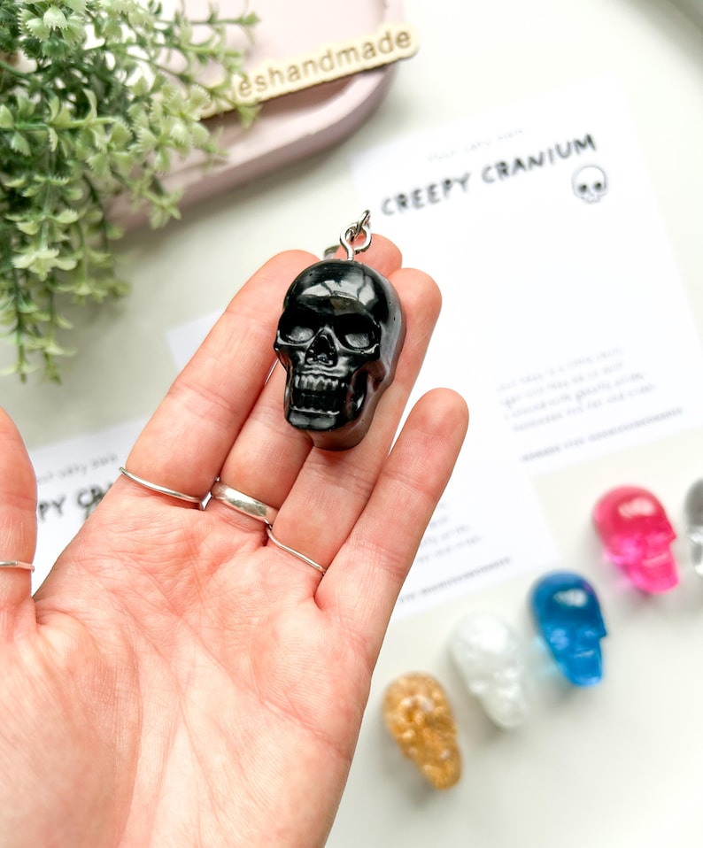 May include: A black skull-shaped keychain is held in a hand, with a silver ring attached. The keychain is glossy and detailed. The text "CREEPY CRANIUM" is visible on a card in the background, along with other colorful skull charms.