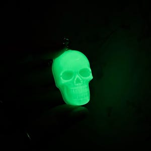 May include: A glowing green skull pendant against a black background. The skull is detailed with eye sockets, a nose cavity, and teeth. The pendant is suspended by a small loop, suggesting it's a charm or accessory.