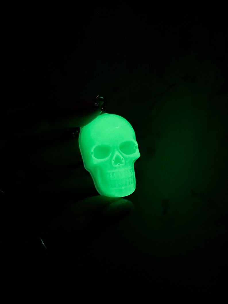 May include: A glowing green skull pendant against a black background. The skull is detailed with eye sockets, a nose cavity, and teeth. The pendant is suspended by a small loop, suggesting it's a charm or accessory.