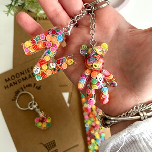 May include: A colourful fruit-themed keychain featuring a letter Z and a bear charm. The keychain is made of clear resin with embedded colourful fruit slices and sprinkles. The keychain is attached to a silver metal chain and ring.