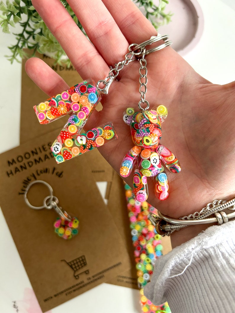 May include: A colourful fruit-themed keychain featuring a letter Z and a bear charm. The keychain is made of clear resin with embedded colourful fruit slices and sprinkles. The keychain is attached to a silver metal chain and ring.