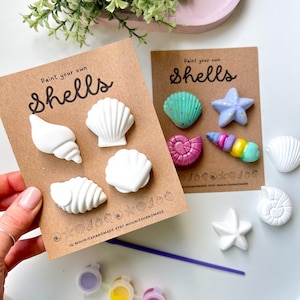 May include: Craft kit featuring paint-your-own shells. The kit includes various shell shapes, paint pots with yellow, pink, and purple paint, and a paintbrush. The text "Paint your own shells" is visible on the packaging.