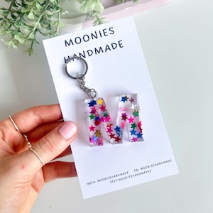 May include: A clear resin keyring in the shape of the letter M, filled with small, multi-coloured star-shaped glitter. The keyring is attached to a silver metal ring and displayed on a white card labelled MOONIES HANDMADE with social media handles.