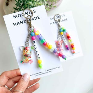 May include: Two handmade keychains with colorful beaded accents. One keychain features a clear bear charm filled with colorful shapes, the other has a pink bear charm. Both keychains have the text "MILLY" and "HOPE" respectively. The keychains are attached to cards that read "MOONIES HANDMADE".