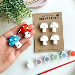 May include: A craft kit featuring mushroom figurines, paint pots, and a paintbrush. Two painted mushrooms, one red and one blue, are held. The kit includes a card with the text "Paint Your Own Mushrooms".