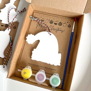 May include: A Christmas ornament craft kit featuring a white ceramic Santa hat, paint pots, a paintbrush, and twine. The kit is presented in a brown cardboard box with the words "Merry Christmas!" printed inside.
