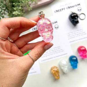 May include: A pink, glitter-filled skull keychain is held up, showcasing its translucent design. The keychain is attached to a silver ring. Other skull keychains in various colors are visible in the background, along with a card that says "Creepy Cranium."