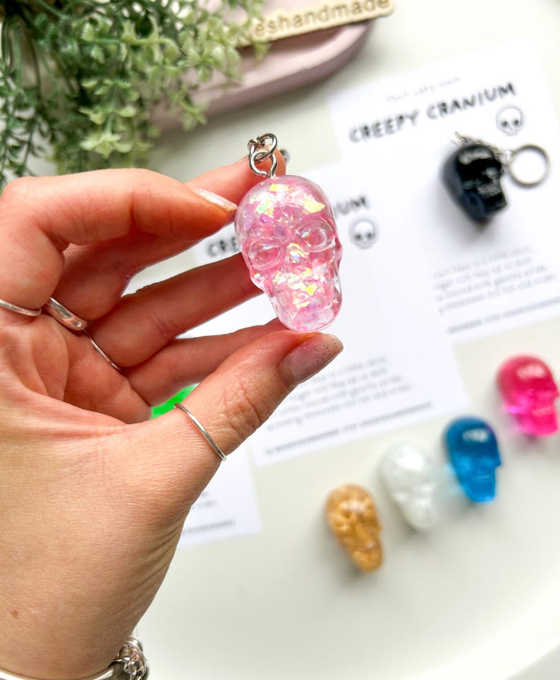 May include: A pink, glitter-filled skull keychain is held up, showcasing its translucent design. The keychain is attached to a silver ring. Other skull keychains in various colors are visible in the background, along with a card that says "Creepy Cranium."