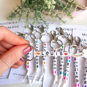 K&ouml;nnte beinhalten: Personalisierte Schl&uuml;sselanh&auml;nger mit Namen, die in wei&szlig;en Buchstabenperlen geschrieben sind. Jeder Schl&uuml;sselanh&auml;nger hat einen anderen Namen, wie LOUIS, MIA und SUMMER, mit bunten Herzakzenten. Die Schl&uuml;sselanh&auml;nger sind an silbernen Ringen befestigt.
