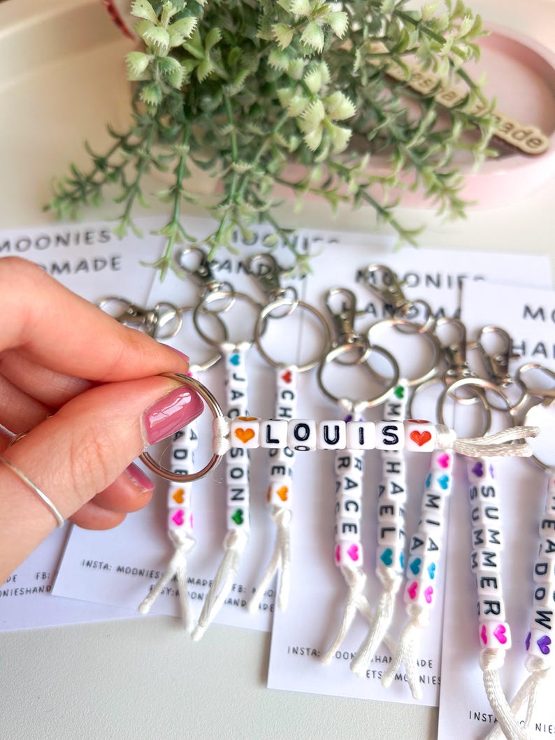 K&ouml;nnte beinhalten: Personalisierte Schl&uuml;sselanh&auml;nger mit Namen, die in wei&szlig;en Buchstabenperlen geschrieben sind. Jeder Schl&uuml;sselanh&auml;nger hat einen anderen Namen, wie LOUIS, MIA und SUMMER, mit bunten Herzakzenten. Die Schl&uuml;sselanh&auml;nger sind an silbernen Ringen befestigt.