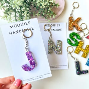 May include: A collection of letter-shaped keychains made of resin with glitter, including a purple J, a silver N, a gold K, a green S, a yellow H, and a dark blue L. Each keychain is attached to a metal ring and displayed on a white card labelled MOONIES HANDMADE.