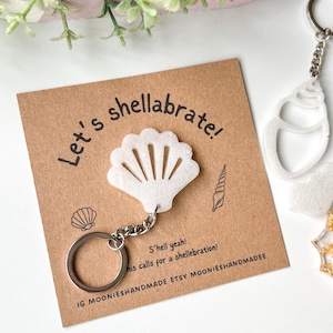 May include: A white seashell keyring with a silver chain. The keyring is attached to a brown card with the text "Let's shellabrate!" and "S'hell yeah! his calls for a shellebration!"