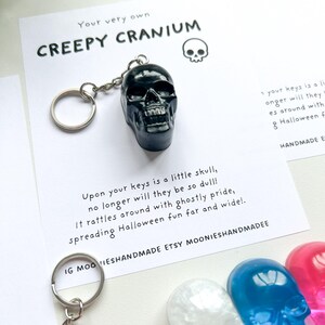 May include: A black skull keychain with a silver keyring. The keychain is displayed on a white card with the text "CREEPY CRANIUM" and a skull illustration. Additional skull keychains in white, blue, and pink are visible.
