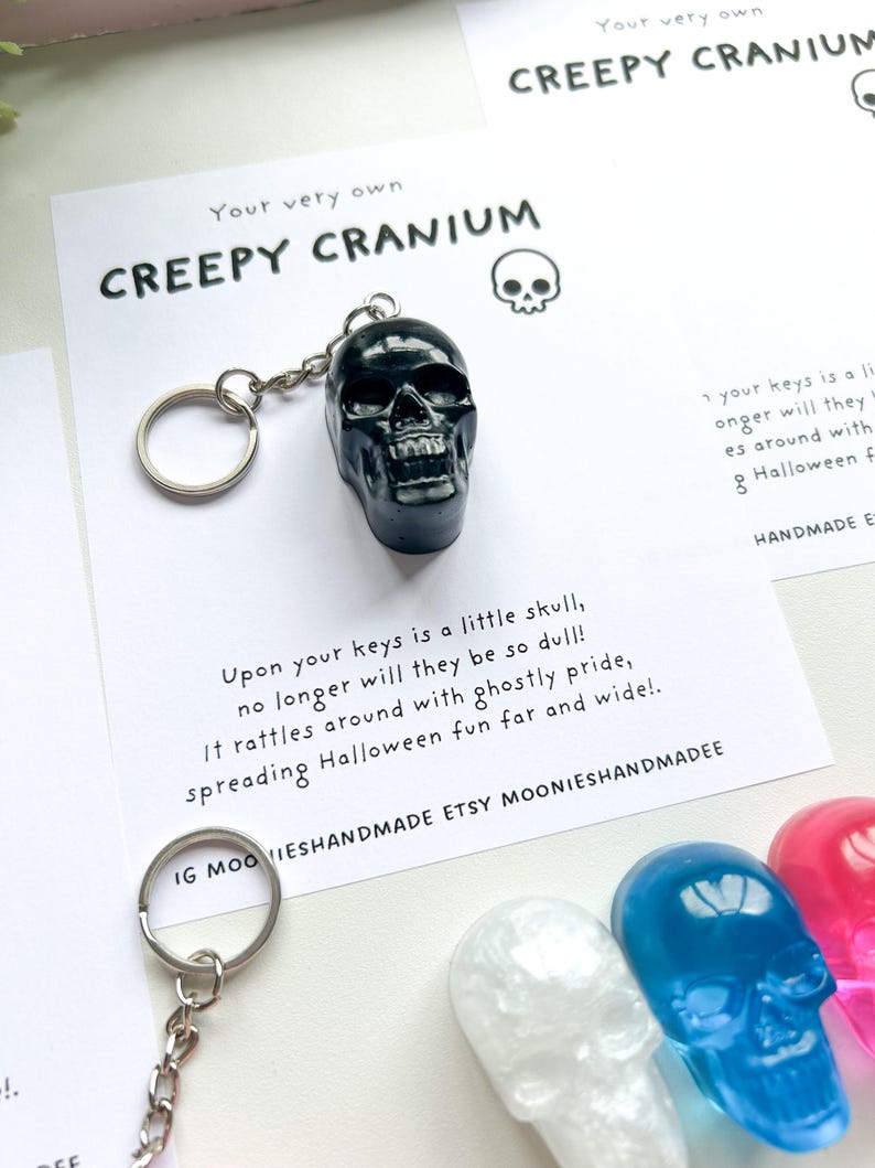 May include: A black skull keychain with a silver keyring. The keychain is displayed on a white card with the text "CREEPY CRANIUM" and a skull illustration. Additional skull keychains in white, blue, and pink are visible.