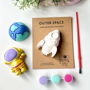 May include: A pottery painting kit with an outer space theme. The kit includes a rocket, astronaut, and planet figurines, paint pots, and a paintbrush. The packaging features the text "Outer Space" and is designed for creative activities.