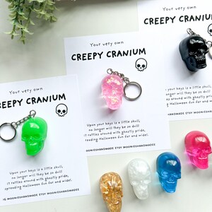 May include: A collection of colorful skull keychains in green, pink, black, gold, white, blue, and fuchsia. Each keychain is paired with a card that reads "Creepy Cranium" and a Halloween-themed message. The keychains are a fun, spooky accessory.