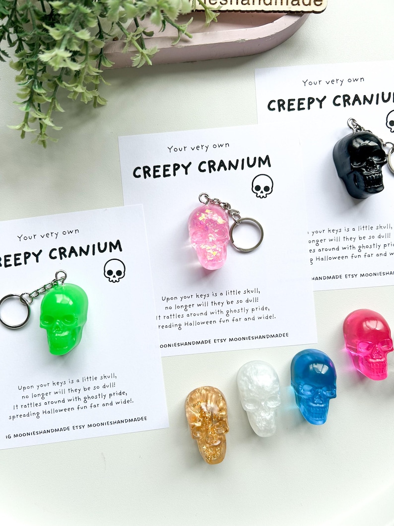 May include: A collection of colorful skull keychains in green, pink, black, gold, white, blue, and fuchsia. Each keychain is paired with a card that reads "Creepy Cranium" and a Halloween-themed message. The keychains are a fun, spooky accessory.